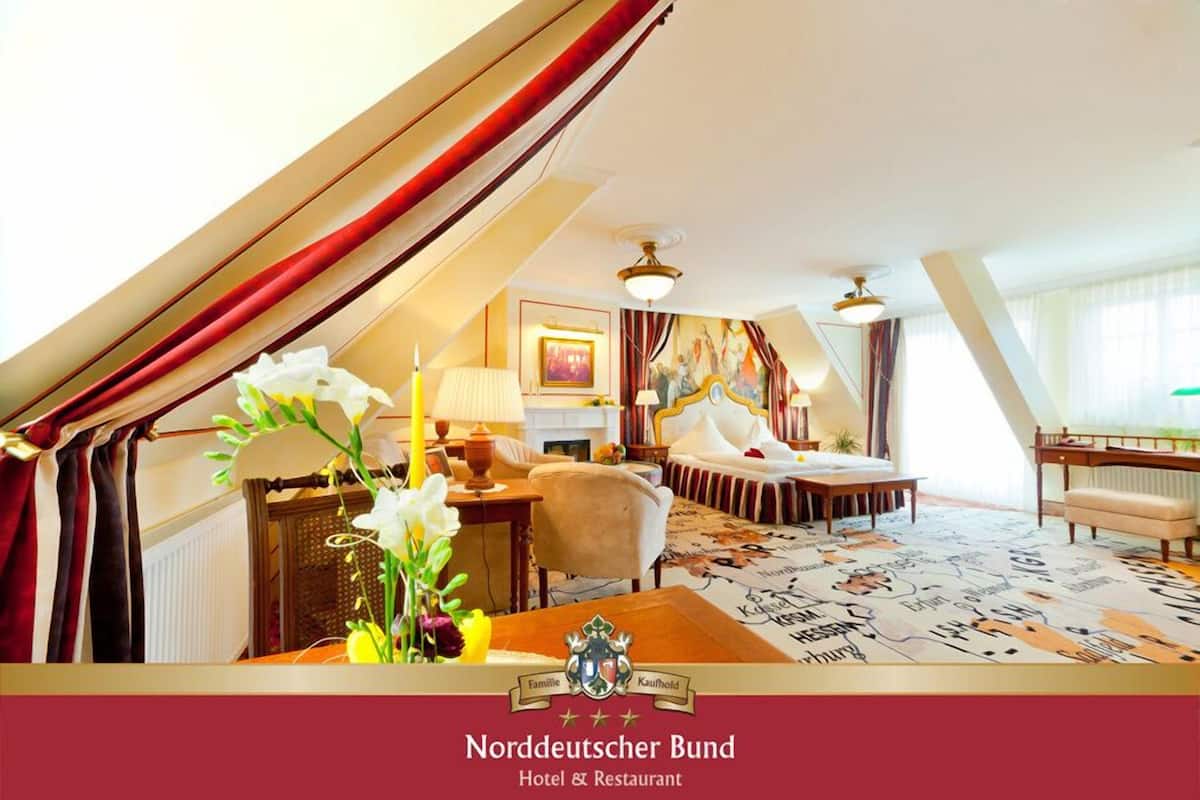 Une chambre d’hôtel avec un plafond en pente, un lit avec une tête de lit blanche, un bureau avec une lampe et un tableau accroché au mur.