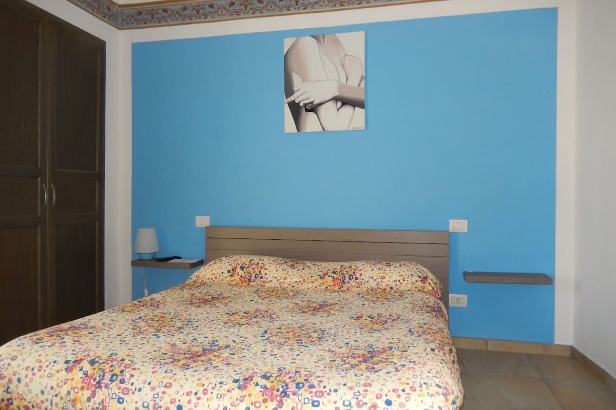 Un dormitorio con pared azul, estructura de cama de madera, cubrecama estampado, mesita de noche con lámpara y un cuadro colgado en la pared.
