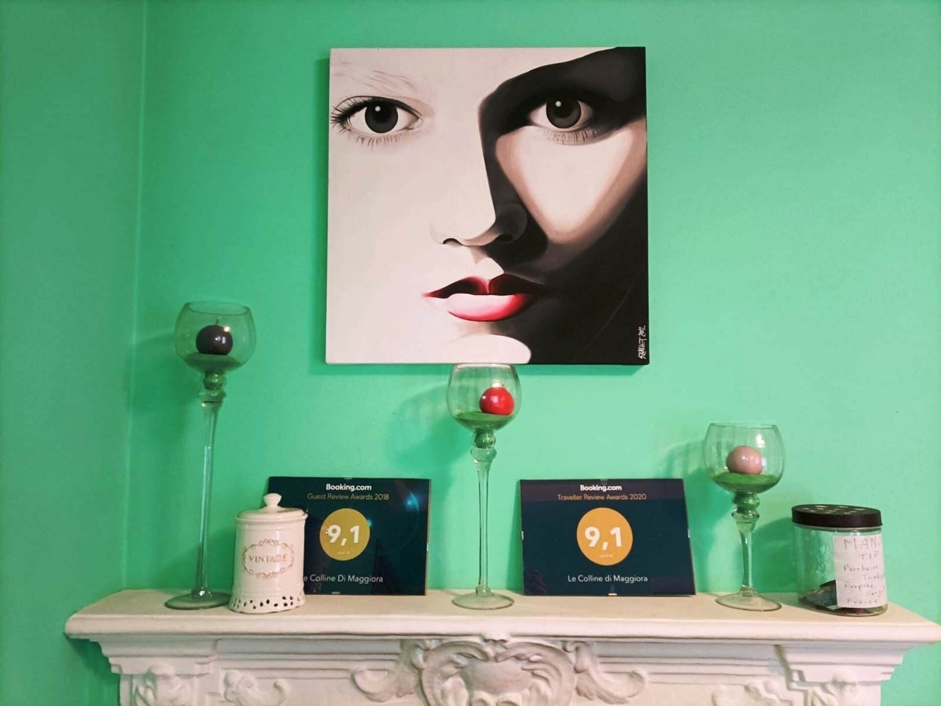 Una pared verde con una obra de arte enmarcada, un manto de chimenea blanco con copas de vino, un tarro antiguo y dos premios Booking.com.