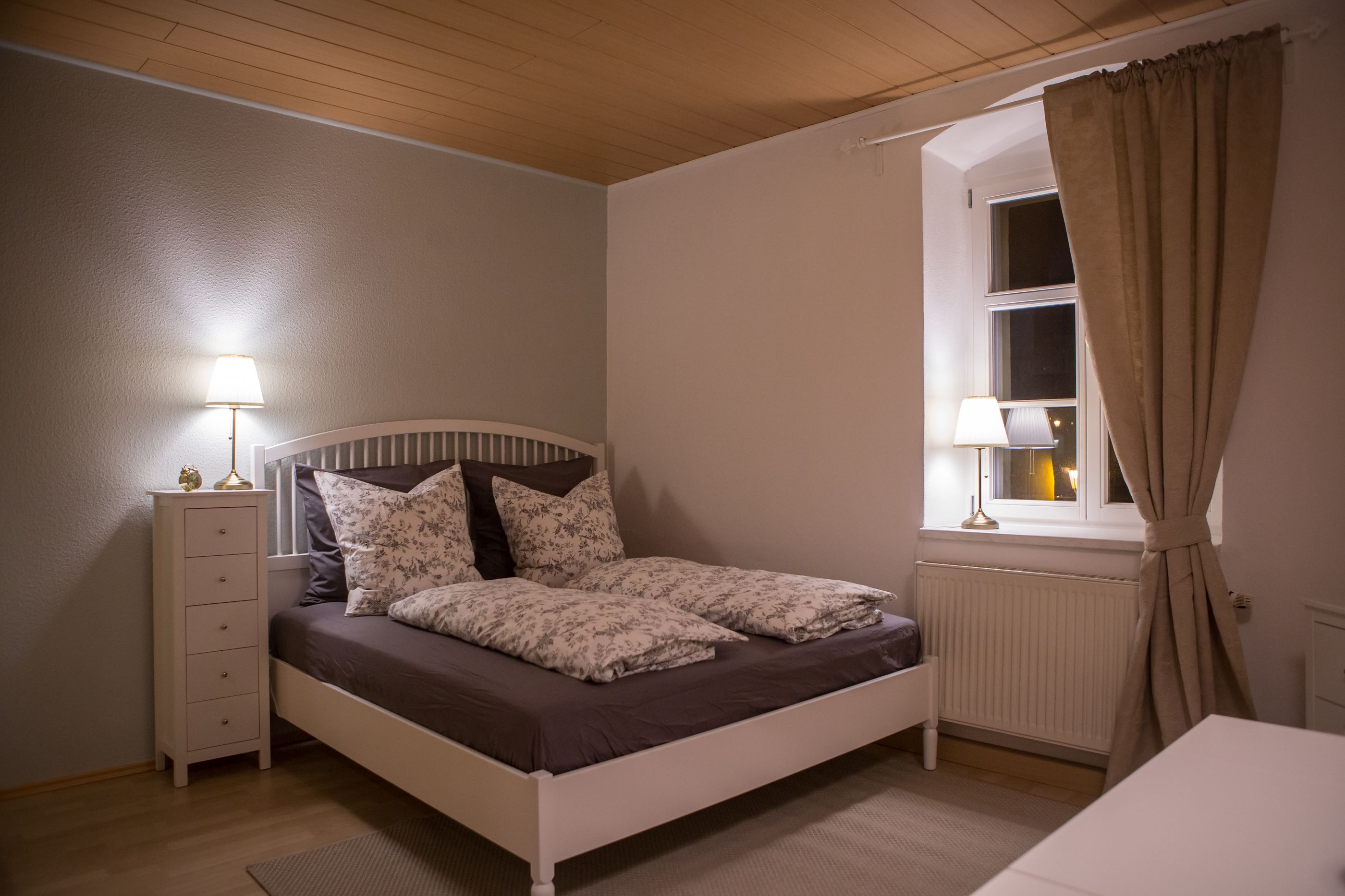 A bedroom with a bed, bedside tables, a window with curtains, and a radiator.
