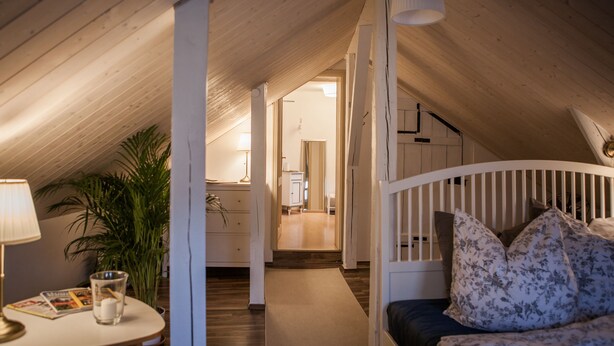 A cozy attic bedroom with a wooden ceiling, a white crib, a bed with floral pillows, a small table with a lamp, and a potted plant. - ApartOne (Bautzen)