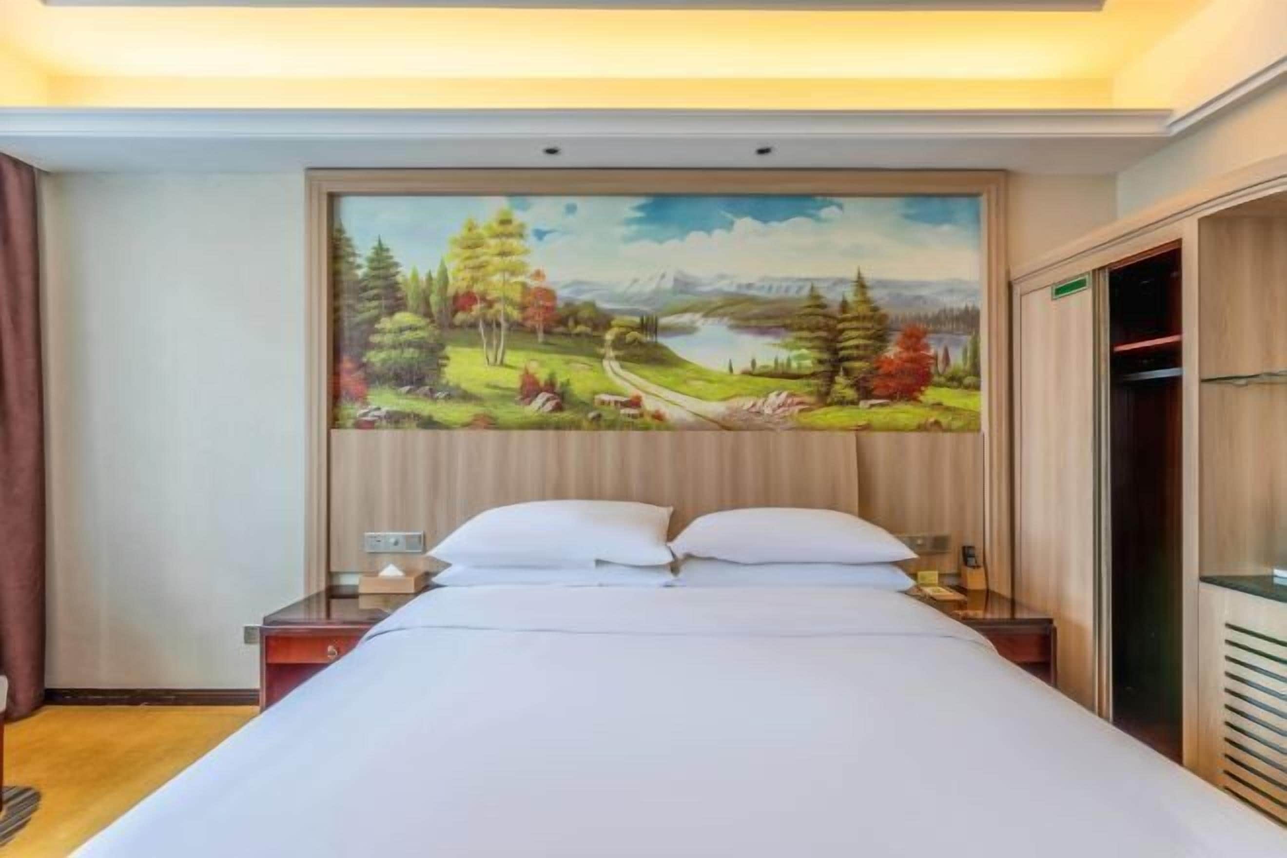 A hotel room with a large bed, a nightstand, a painting of a landscape, and a wardrobe.