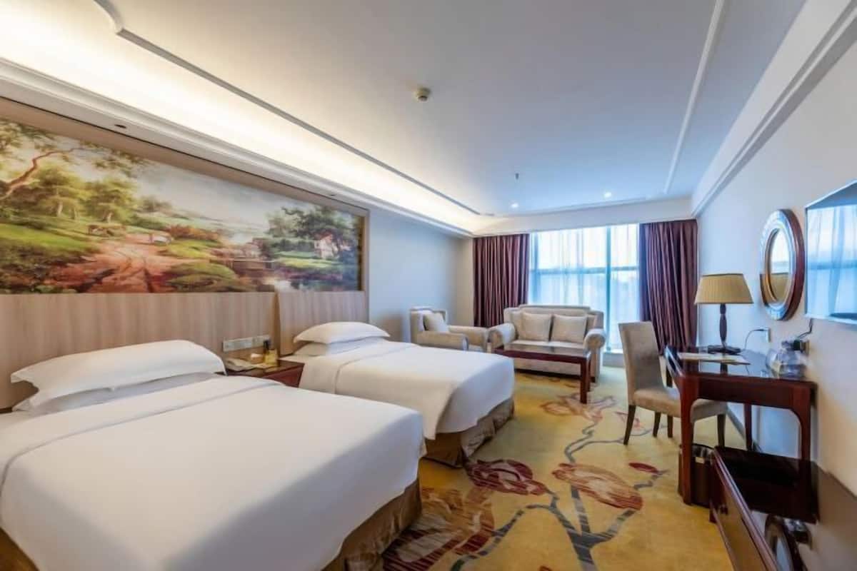 A hotel room with two beds, a desk, a chair, a large painting, and a view of the outdoors.
