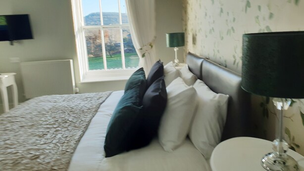 A bedroom with a bed, a nightstand, a lamp, a TV on the wall, and a window with curtains. - Orles Barn (Ross-on-Wye)