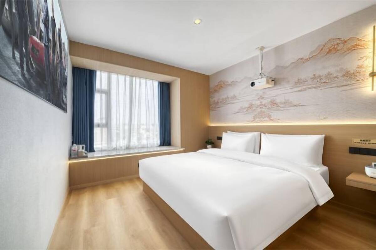 A hotel room with a large bed, a window with curtains, a wall-mounted projector, and a wooden floor.