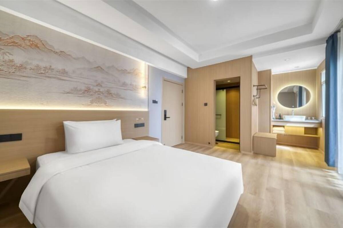 A hotel room with a large bed, a wall-mounted artwork, a bathroom with a mirror, and a wooden floor.