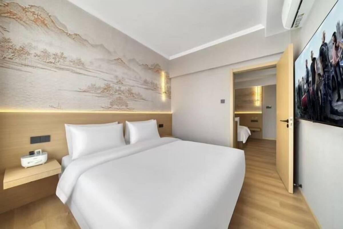 A modern hotel room with a large bed, a nightstand, a wall-mounted air conditioner, and a painting on the wall.