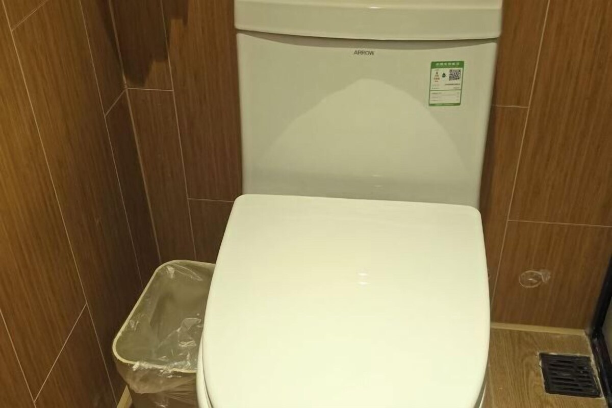 Et hvidt toilet med en grøn klistermærke, en plastik skraldespand og trævægge.