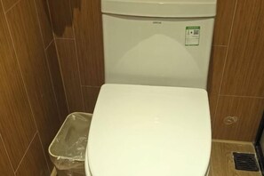Et hvidt toilet med en grøn klistermærke, en plastik skraldespand og trævægge.