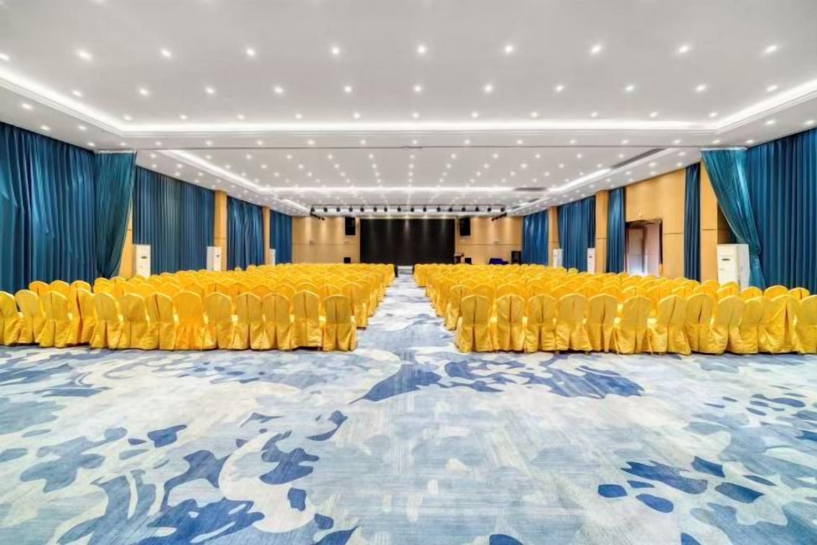 A large conference room with rows of yellow chairs, a stage, and a blue carpet.