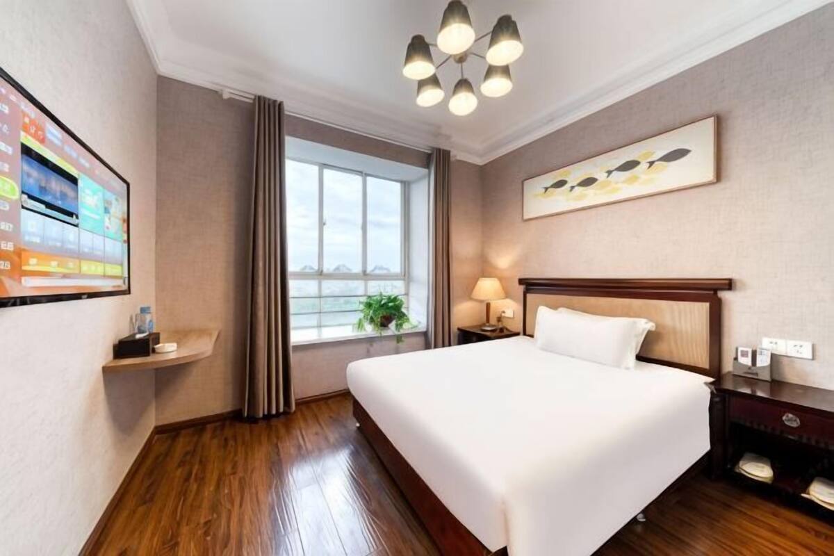 A hotel room with a large bed, a window with curtains, a wall-mounted TV, and a painting.