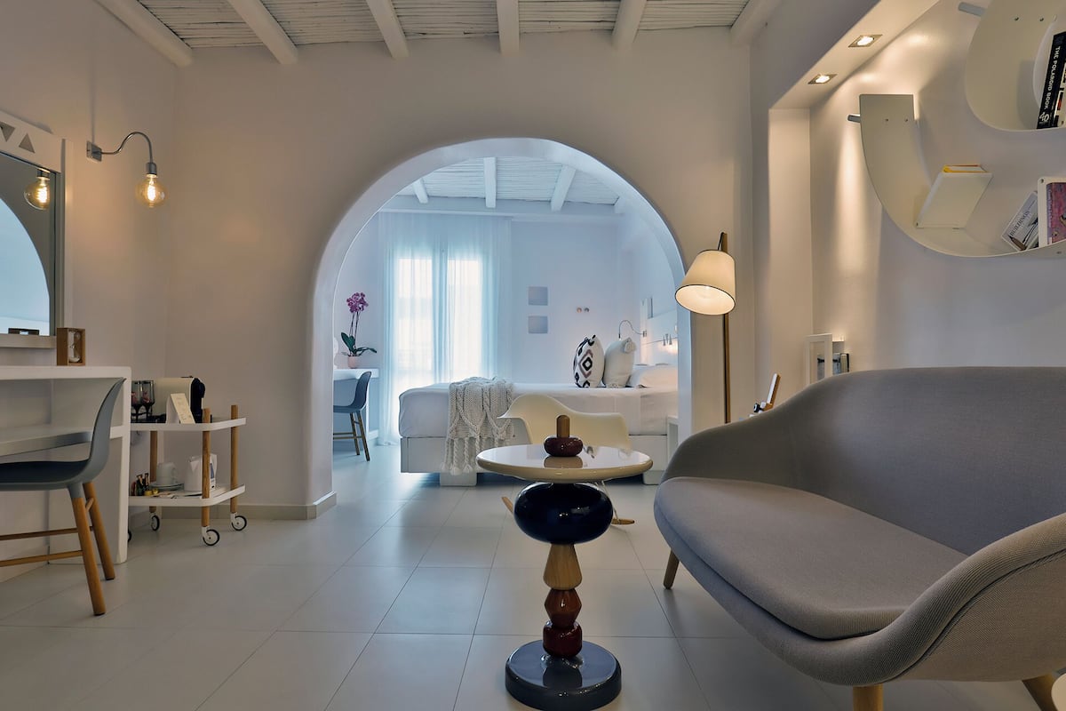 Una sala de estar moderna y minimalista con un sofá, una mesa de centro y un escritorio con una silla. Un arco de entrada conduce a un dormitorio con una cama y un escritorio.