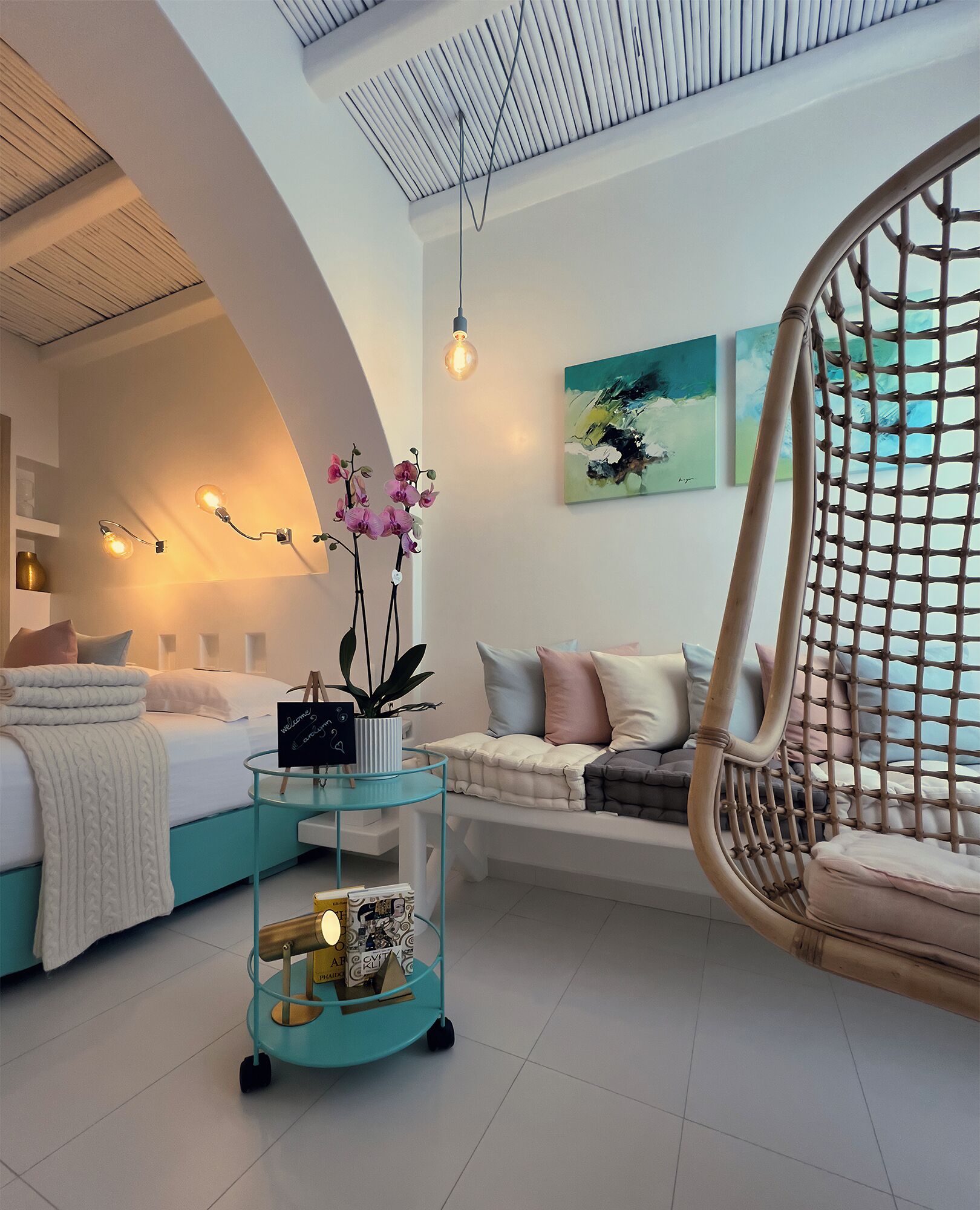 A modern hotel room with a bed, a sofa, a wicker chair, a glass coffee table, and artwork on the wall.