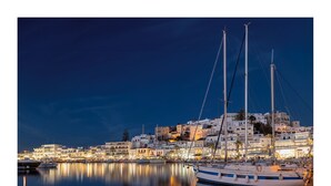 Una marina con un velero amarrado, agua tranquila que refleja edificios, y un paisaje urbano nocturno al fondo.