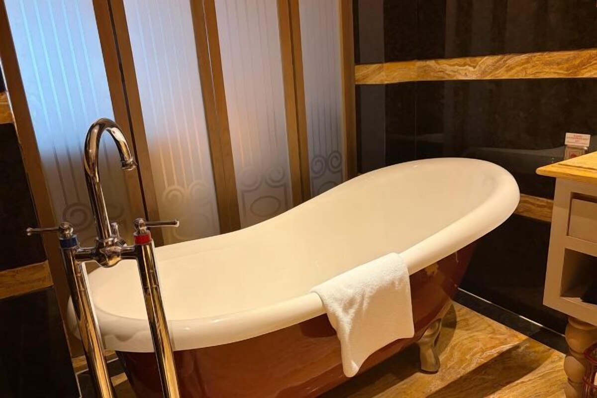 A freestanding bathtub in a bathroom with a wooden floor and a window with blinds.