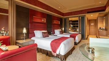 Habitación de hotel con dos camas, un área de descanso con una silla roja, una mesa de vidrio y un baño visible a través de una puerta abierta.