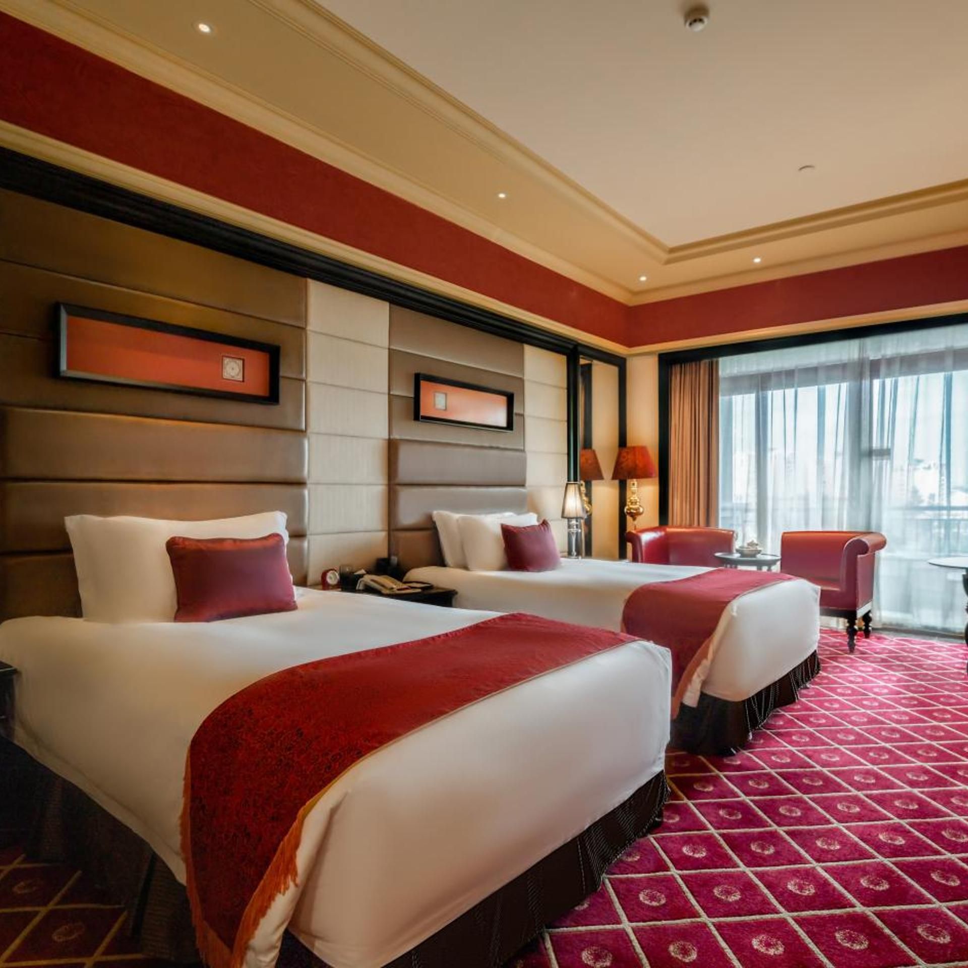 A hotel room with two beds, a red carpet, a large window with curtains, and a view of the city.