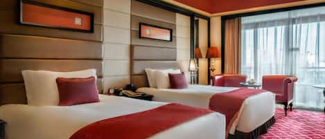 A hotel room with two beds, a red carpet, a large window with curtains, and a view of the city.