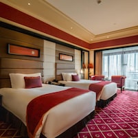 Una habitación de hotel con dos camas, alfombra roja, un ventanal con cortinas y vistas a la ciudad.