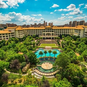 A hotel with a central pool, surrounded by lush greenery and a city skyline in the background.