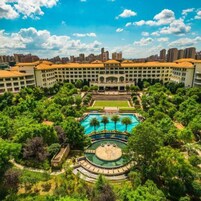 Un hotel con piscina central, rodeado de exuberante vegetación y un horizonte urbano de fondo.