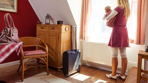 Une pièce avec un meuble en bois, une chaise en osier, une valise et une personne tenant un bébé près d’une fenêtre.