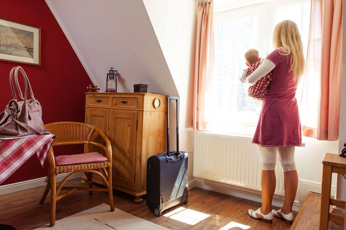 A room with a wooden cabinet, a wicker chair, a suitcase, and a person holding a baby by a window.