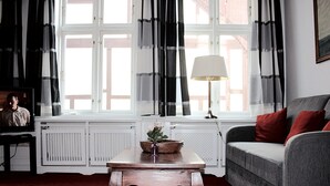 Un salon avec une table basse en bois, un canapé gris agrémenté de coussins rouges, une lampe et une fenêtre avec des rideaux.
