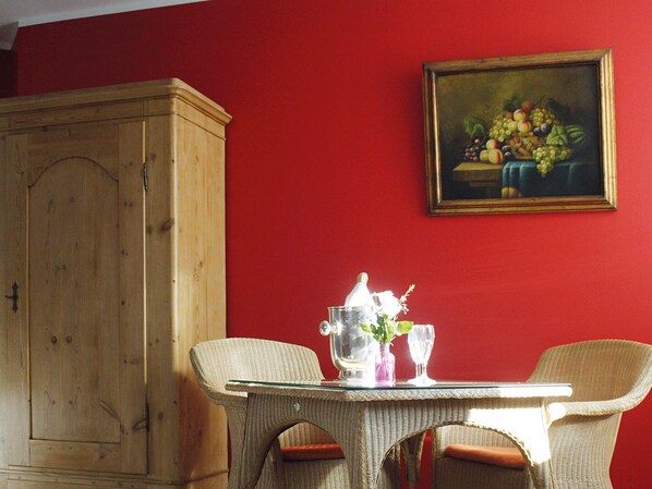 A dining area with wicker chairs, a glass table, and a painting of fruit on the wall.