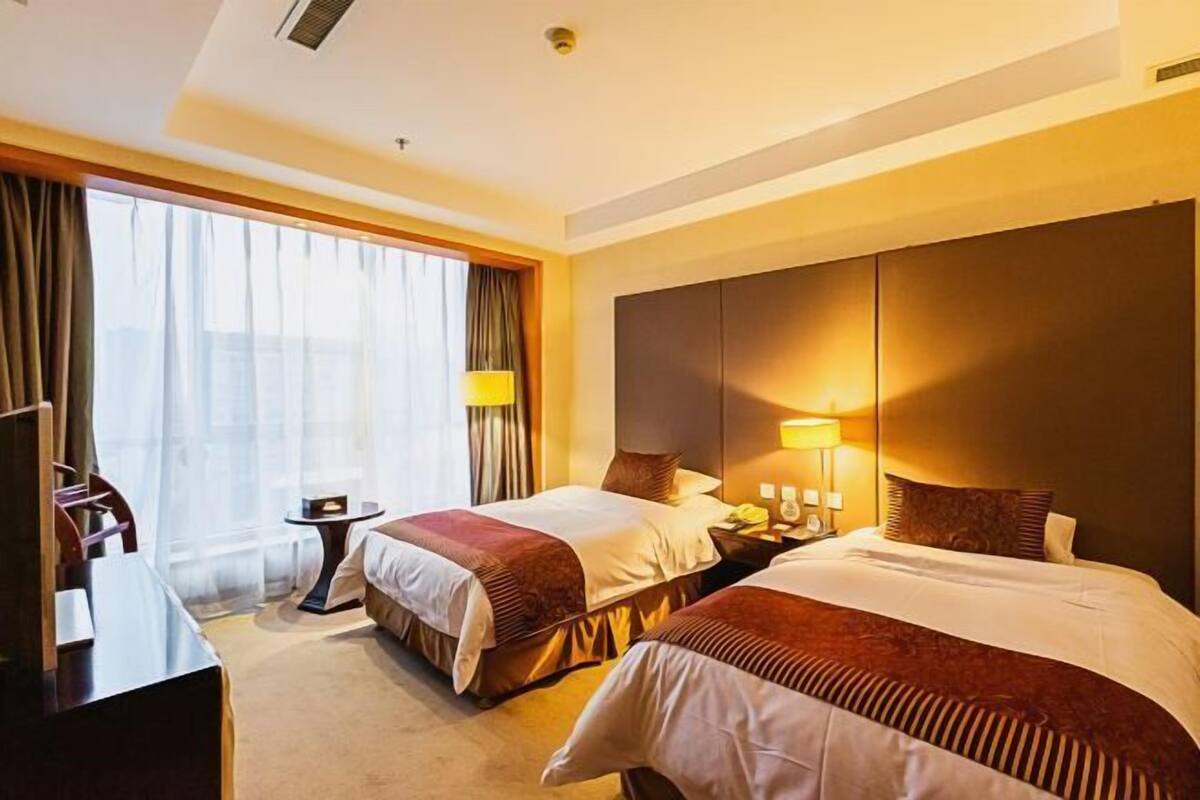 A hotel room with two beds, a desk, a chair, a lamp, and a large window with curtains.