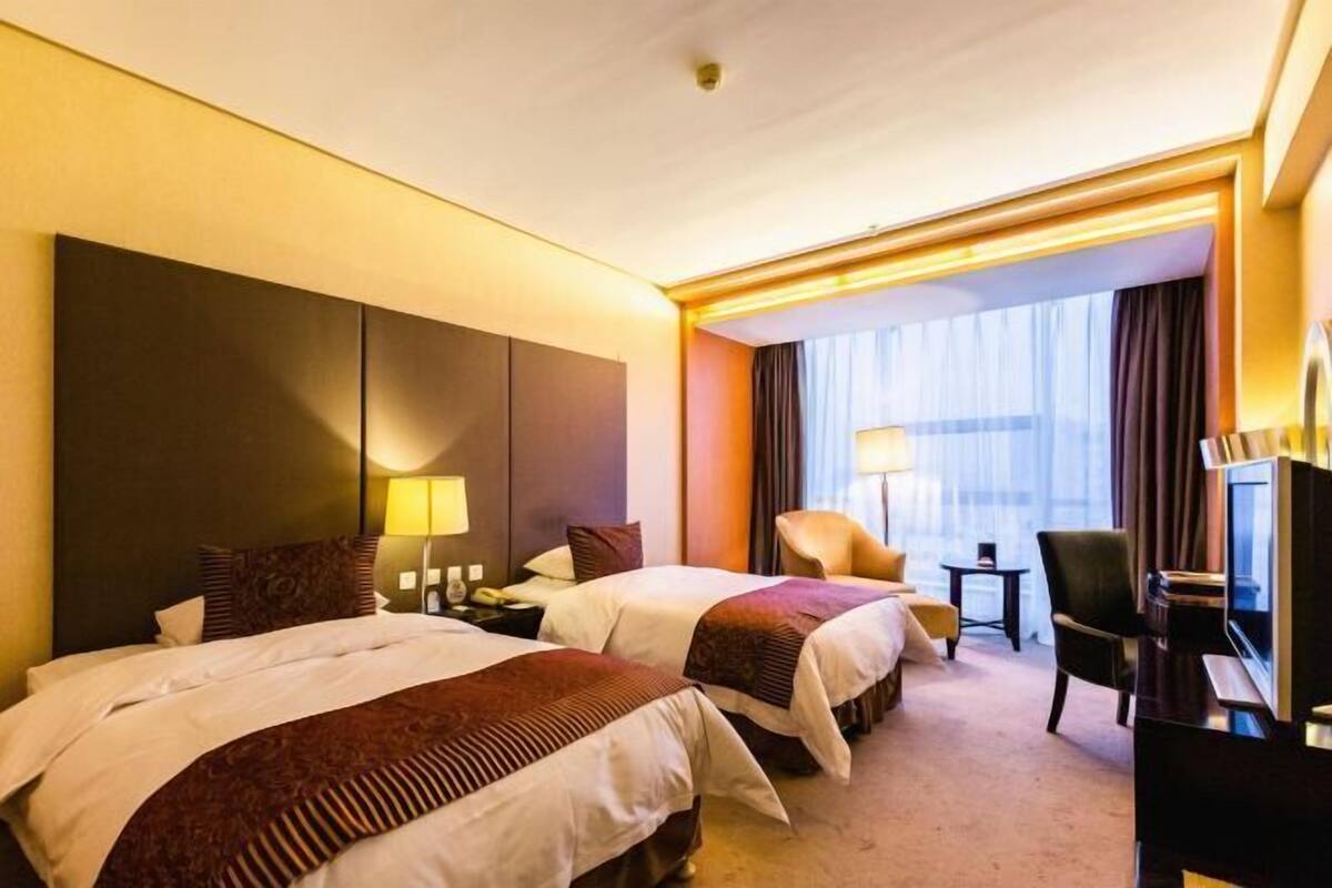 A hotel room with two beds, a desk, a chair, a TV, and a large window with curtains.