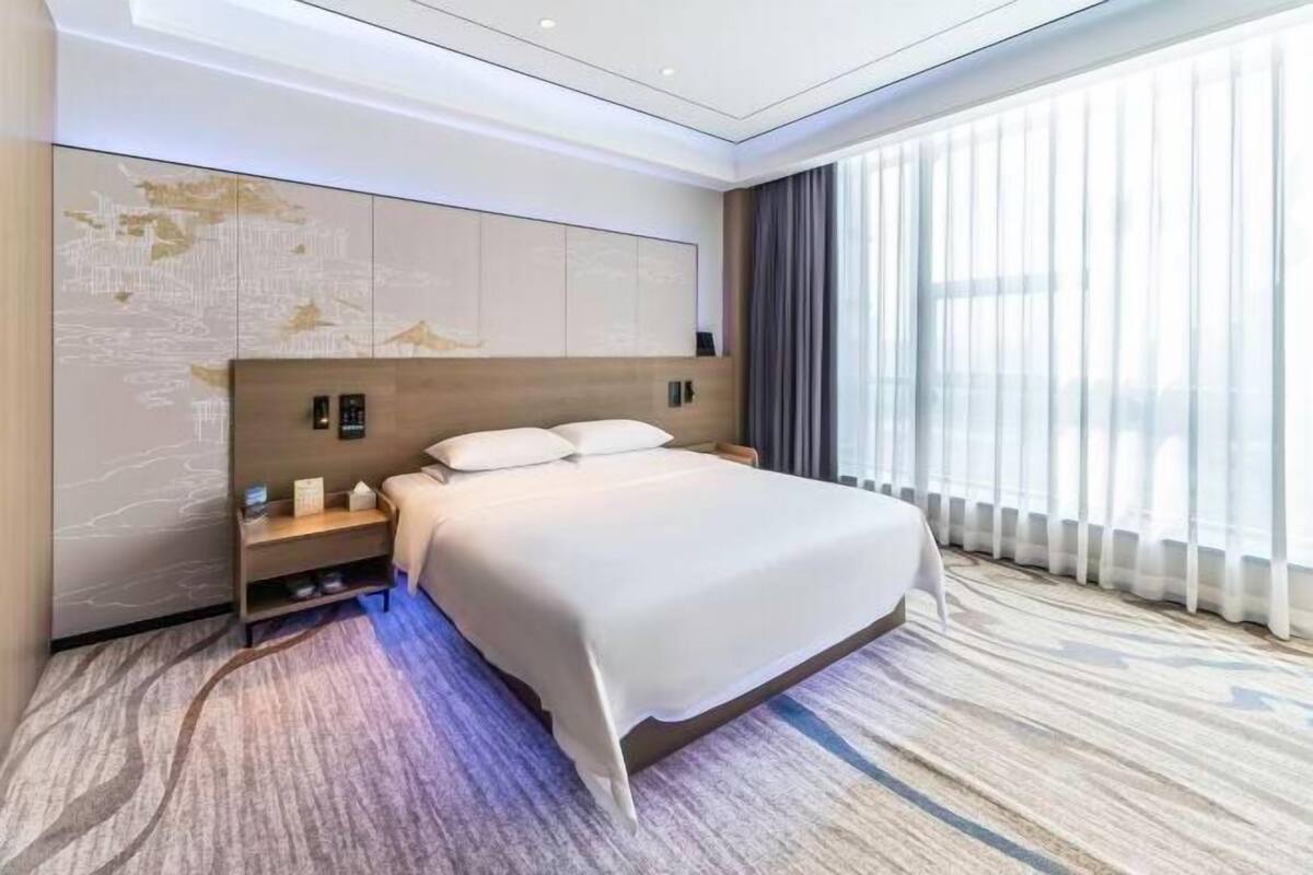 A modern hotel room with a large bed, a headboard with built-in controls, a bedside table with a lamp, and a large window with sheer curtains.