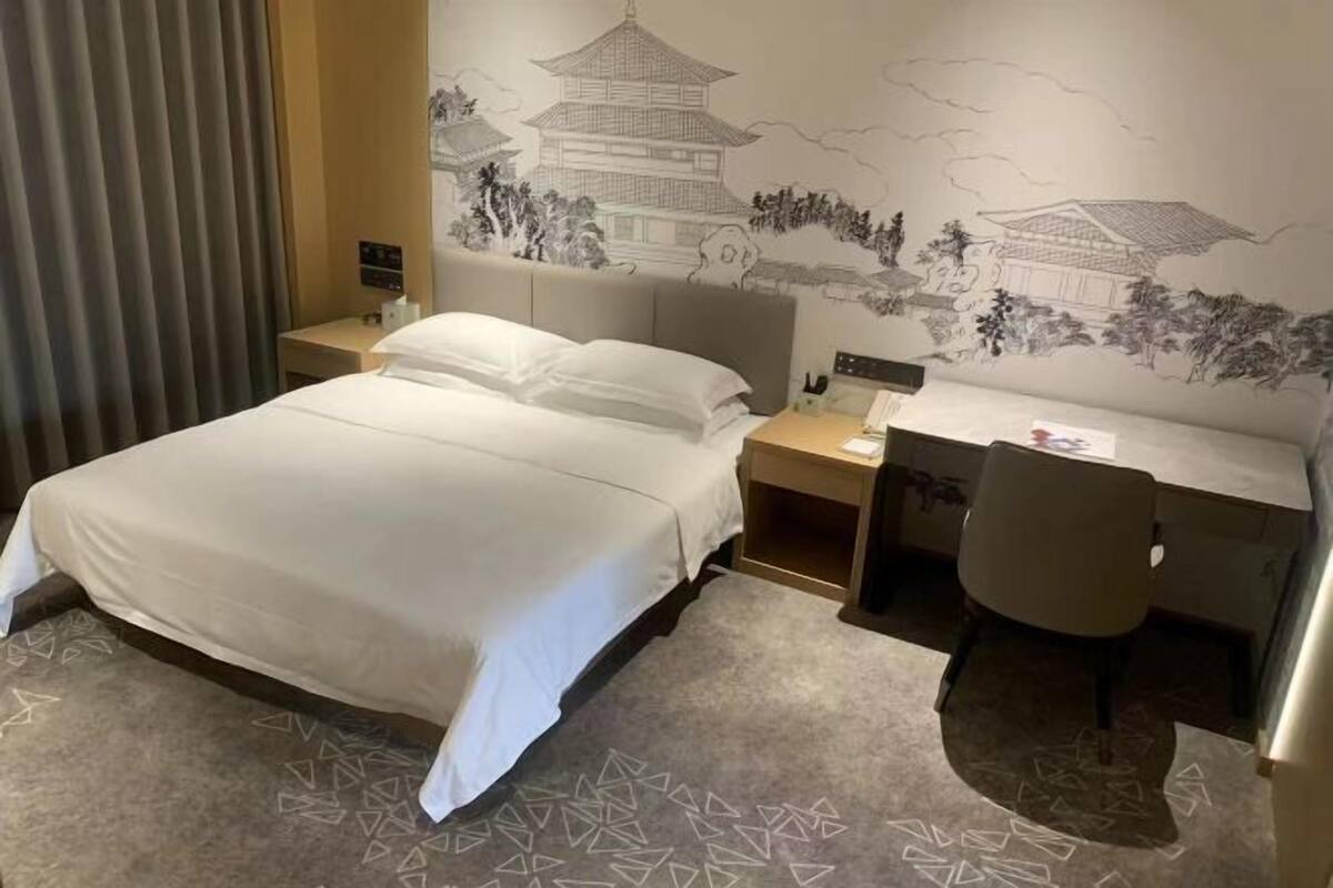 A hotel room with a bed, a desk, and a chair, a nightstand, and a wall with traditional artwork.
