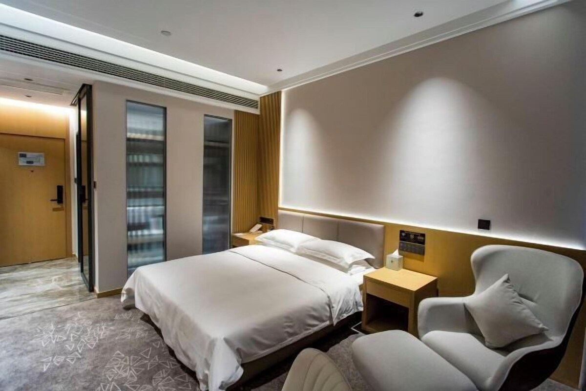 A modern hotel room with a large bed, a sitting area with a chair, a bedside table, and a wall-mounted TV.
