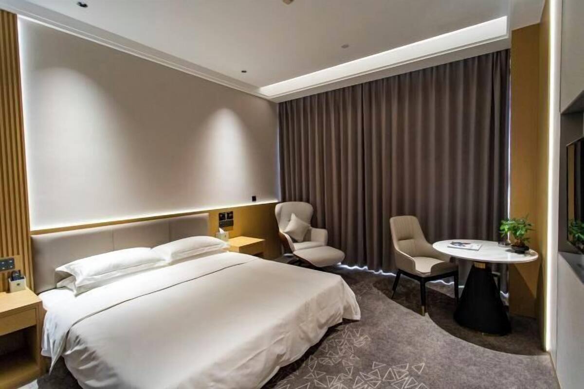 A hotel room with a bed, two armchairs, a small table, a plant, and a large window with curtains.