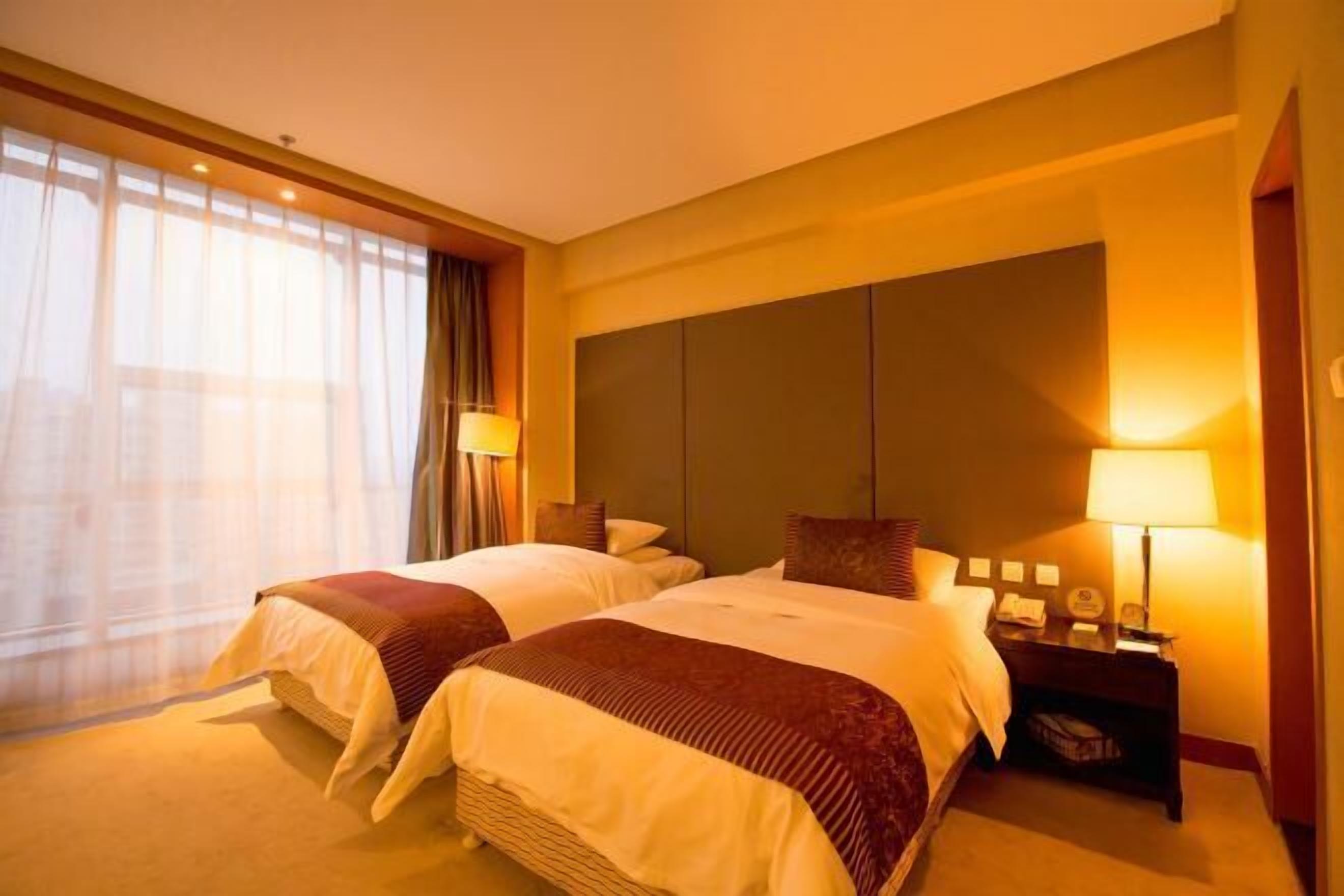 A hotel room with two beds, a large window with curtains, a nightstand with a lamp, and a wall-mounted panel.