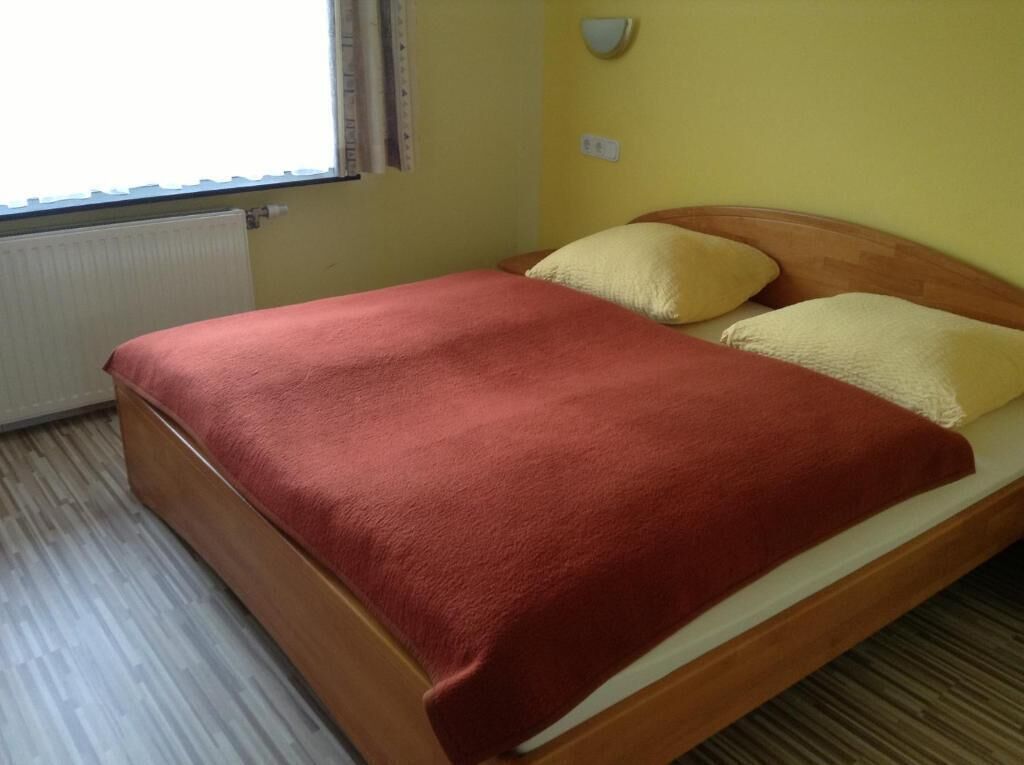 Una cama individual con estructura de madera, una colcha roja y dos almohadas.