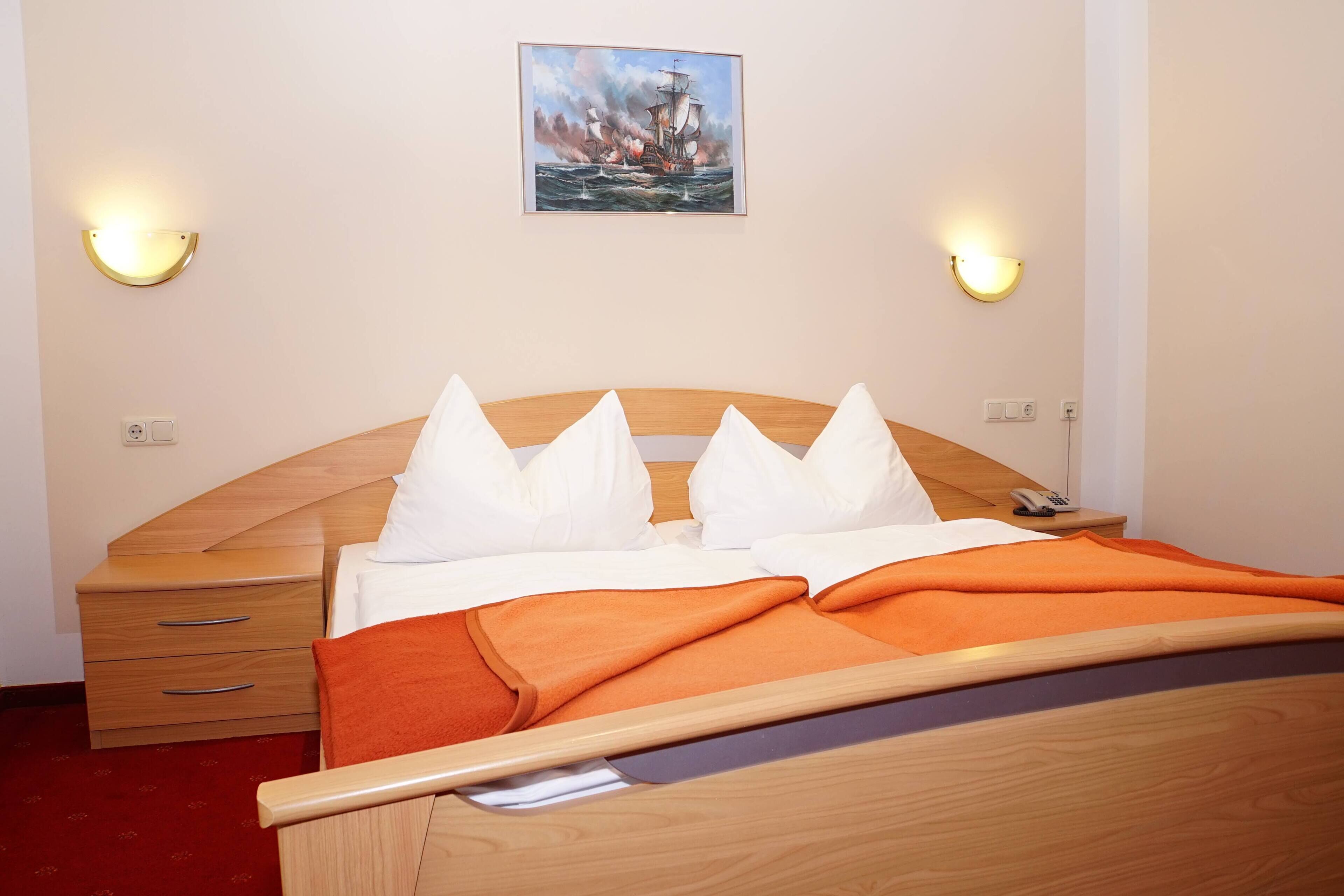 Habitación de hotel con cama de madera, almohadas blancas, manta naranja, mesita de noche con teléfono y un cuadro de un barco en la pared.
