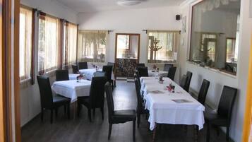 Un comedor con varias mesas y sillas, ventanas amplias con cortinas y vista al exterior.