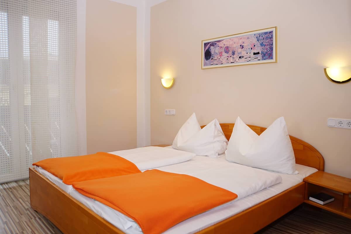 Habitación de hotel con una cama de madera, ropa de cama blanca y una manta anaranjada. Hay dos lámparas de pared y un cuadro en la pared.