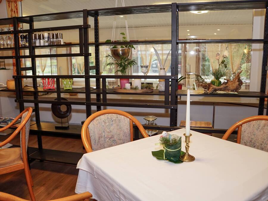 A dining area with a table set for two, surrounded by wooden chairs. A large window with a view of greenery is visible behind a black metal partition.