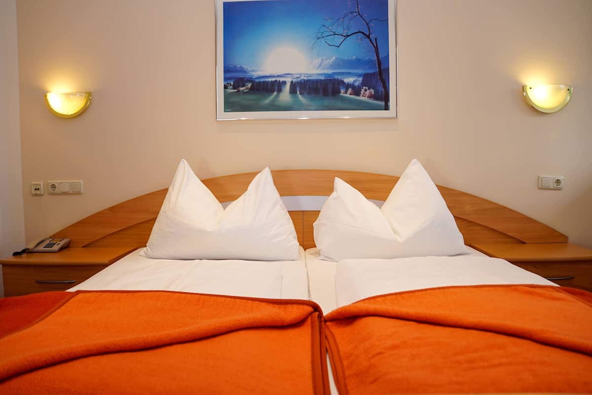 Una habitación de hotel con una cama doble, dos almohadas, un edredón naranja, una mesita de noche con un teléfono y un cuadro en la pared.