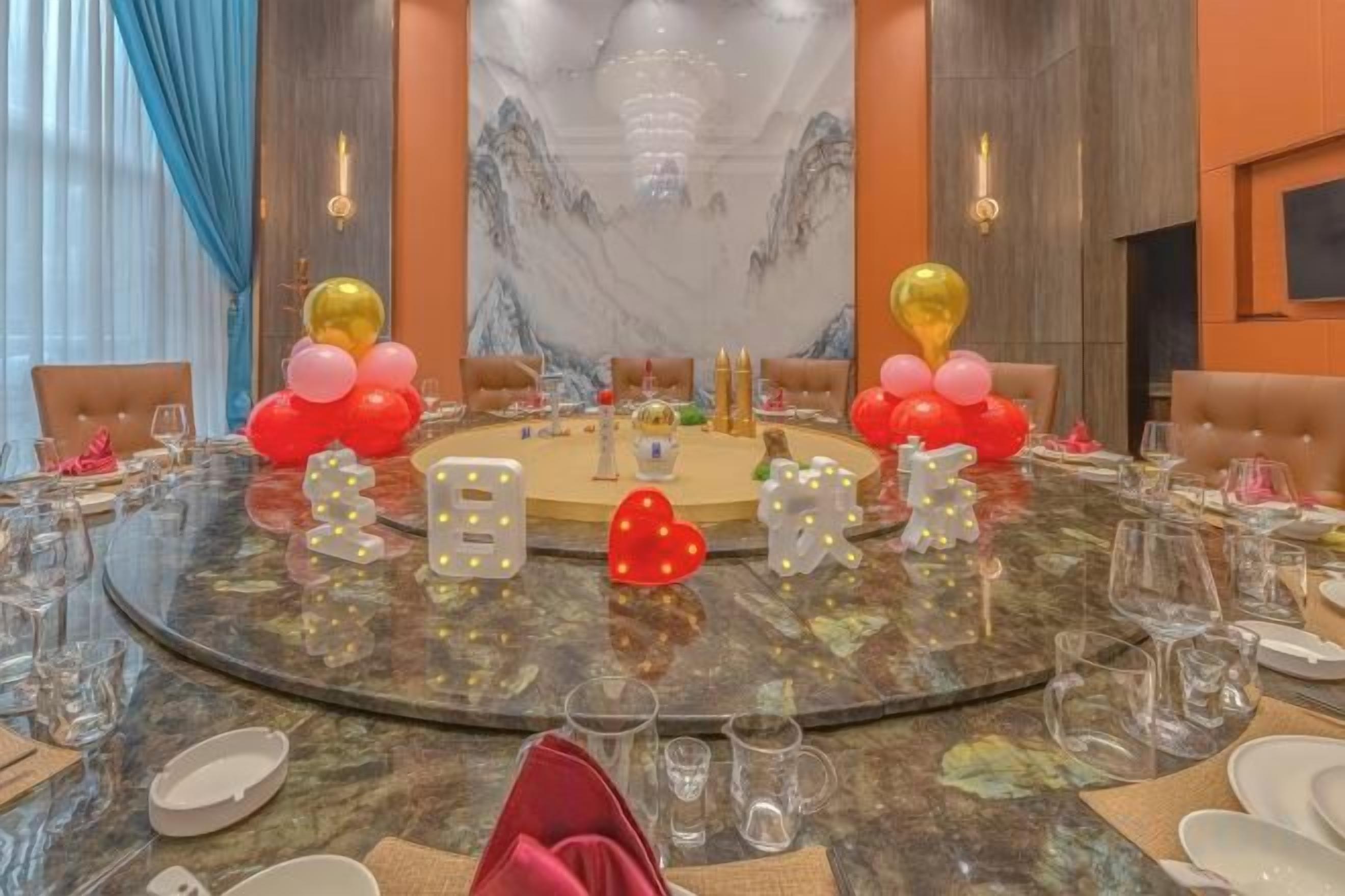Une table ronde dressée pour un événement officiel, entourée de chaises, avec un lustre au-dessus et un grand tableau accroché au mur.