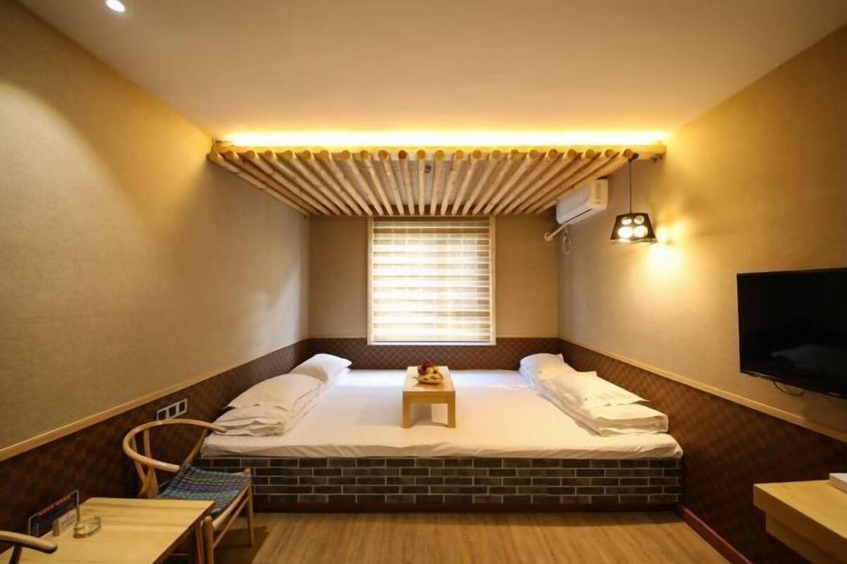 Ein kleines, gemütliches Hotelzimmer mit einem Einzelbett, einem kleinen Tisch, einem Sessel, einem Fernseher und einem Fenster mit Jalousien.