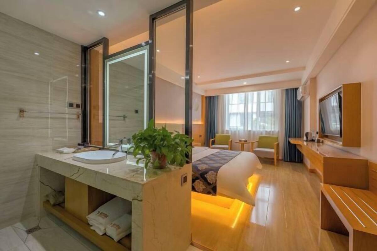 Ein modernes Hotelzimmer mit großem Spiegel, einem Bett mit Kopfteil, einem Nachttisch, einem Fernseher und einem Badezimmer mit Waschbecken und Spiegel.