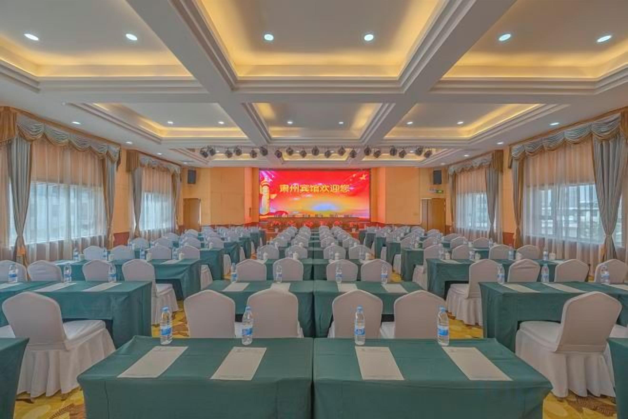 A conference room with rows of tables and chairs, a stage with a screen, and a ceiling with recessed lighting.