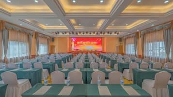 A conference room with rows of tables and chairs, a stage with a screen, and a ceiling with recessed lighting.