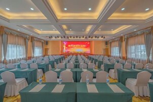 Un salón de conferencias con filas de mesas y sillas, un escenario con una pantalla y un techo con iluminación empotrada.