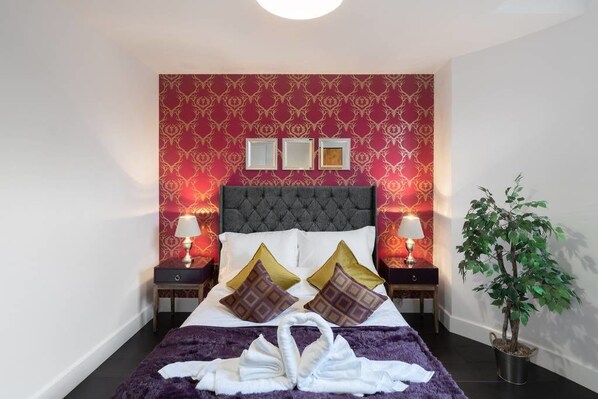 A neatly made bed with a tufted headboard, two bedside tables with lamps, a wall-mounted picture frame, and a potted plant.