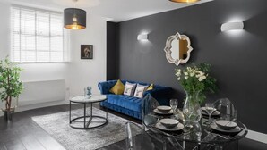 Ein modernes Wohnzimmer mit dunkler Wand, einem blauen Sofa, einem runden Esstisch für sechs Personen und einem großen Fenster mit Jalousien.